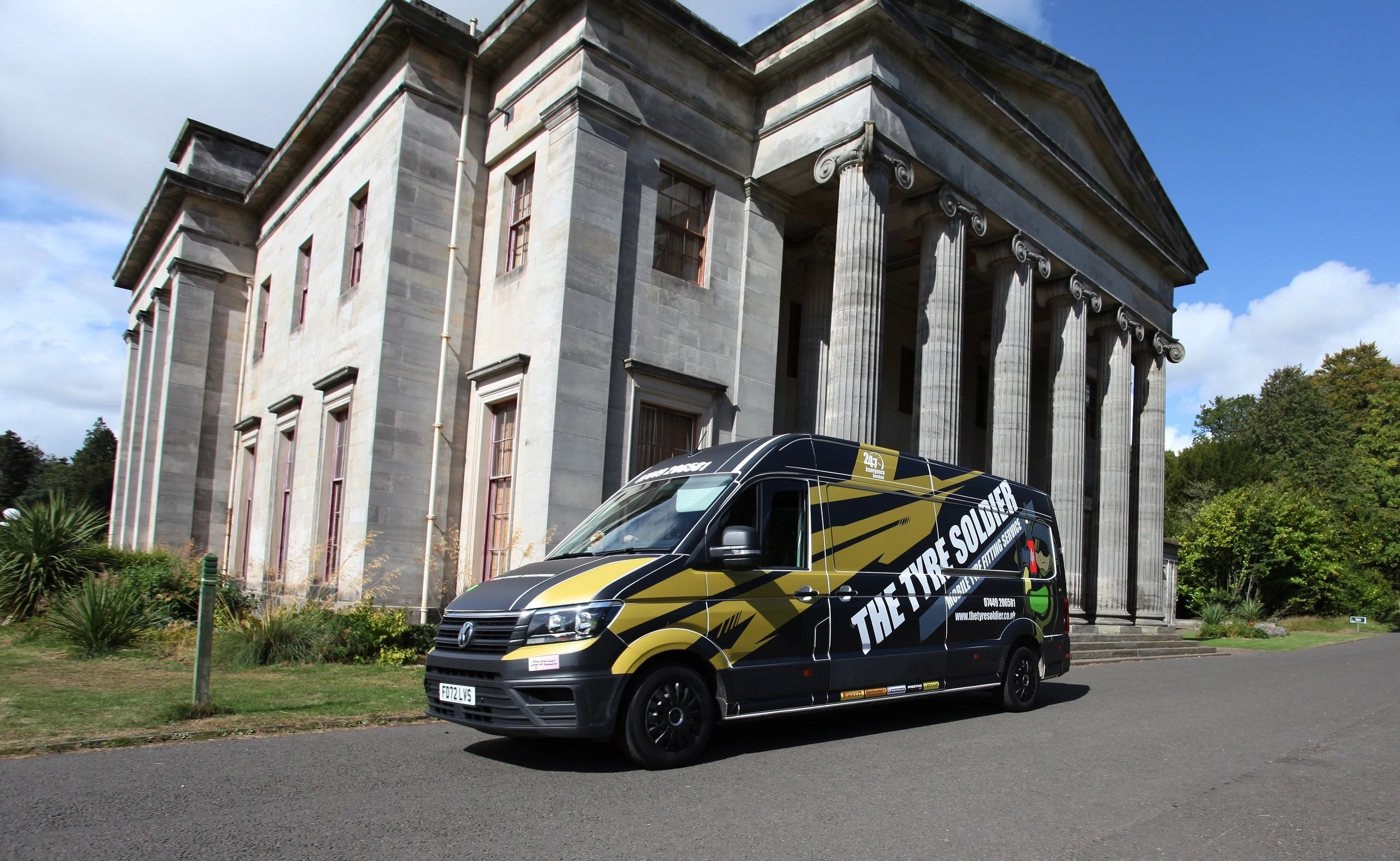 The Tyre Soldier black VW Crafter van with bright military-gold Saltire stripes parked in front of a grand columned Georgian estate building.